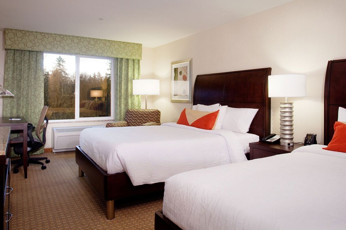 A hotel room with two beds, a desk with a computer, a chair, a TV, and a window with curtains.