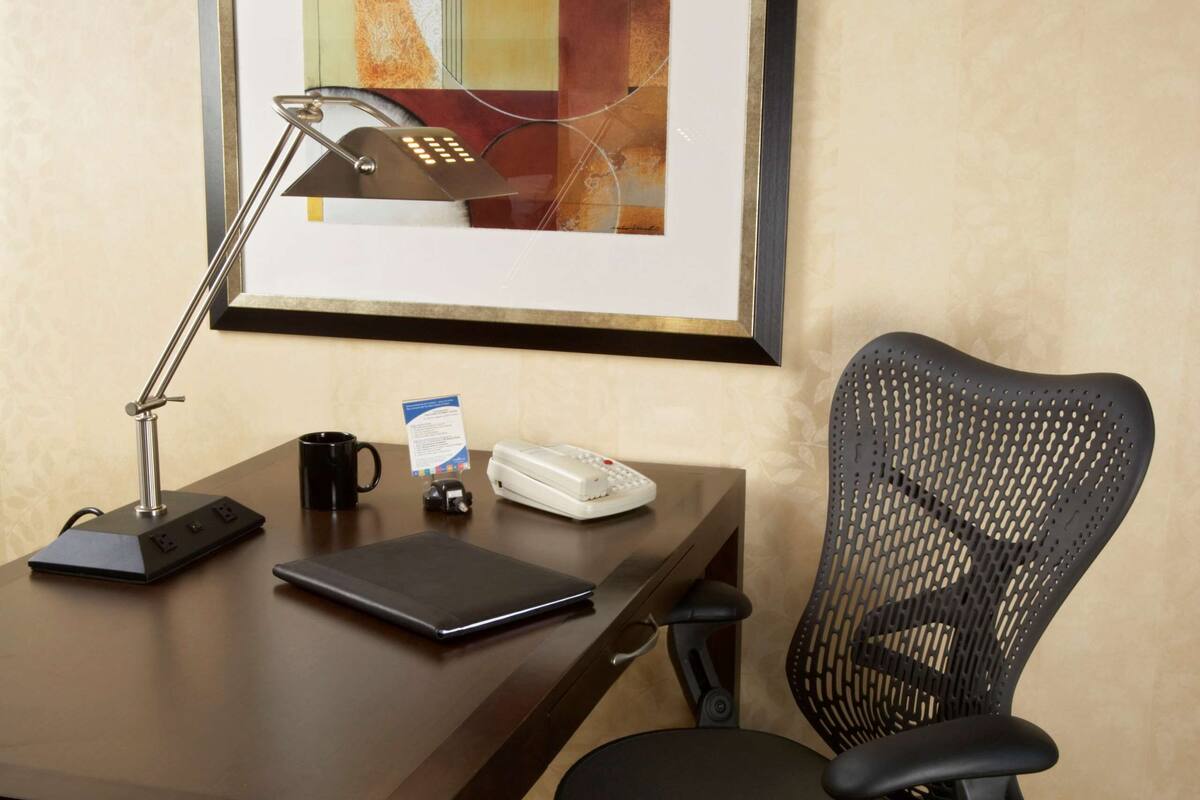 A desk with a laptop, a phone, a coffee mug, a framed abstract painting, and a black office chair.