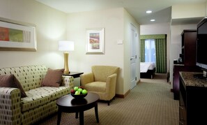 A hotel room with a sofa, armchair, dining table with fruit, a lamp, a painting, a bed, and a TV.
