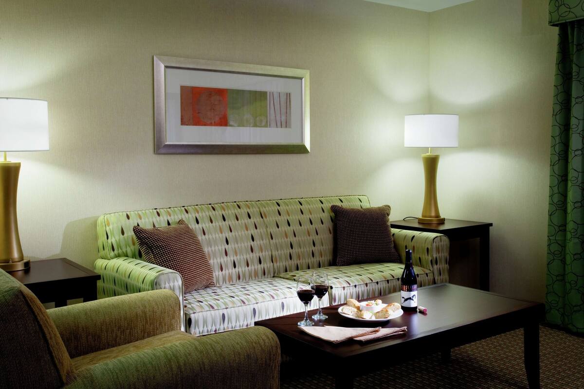 A living room with a sofa, two armchairs, a coffee table, and two lamps. There is a framed picture on the wall and a bottle of wine on the table.