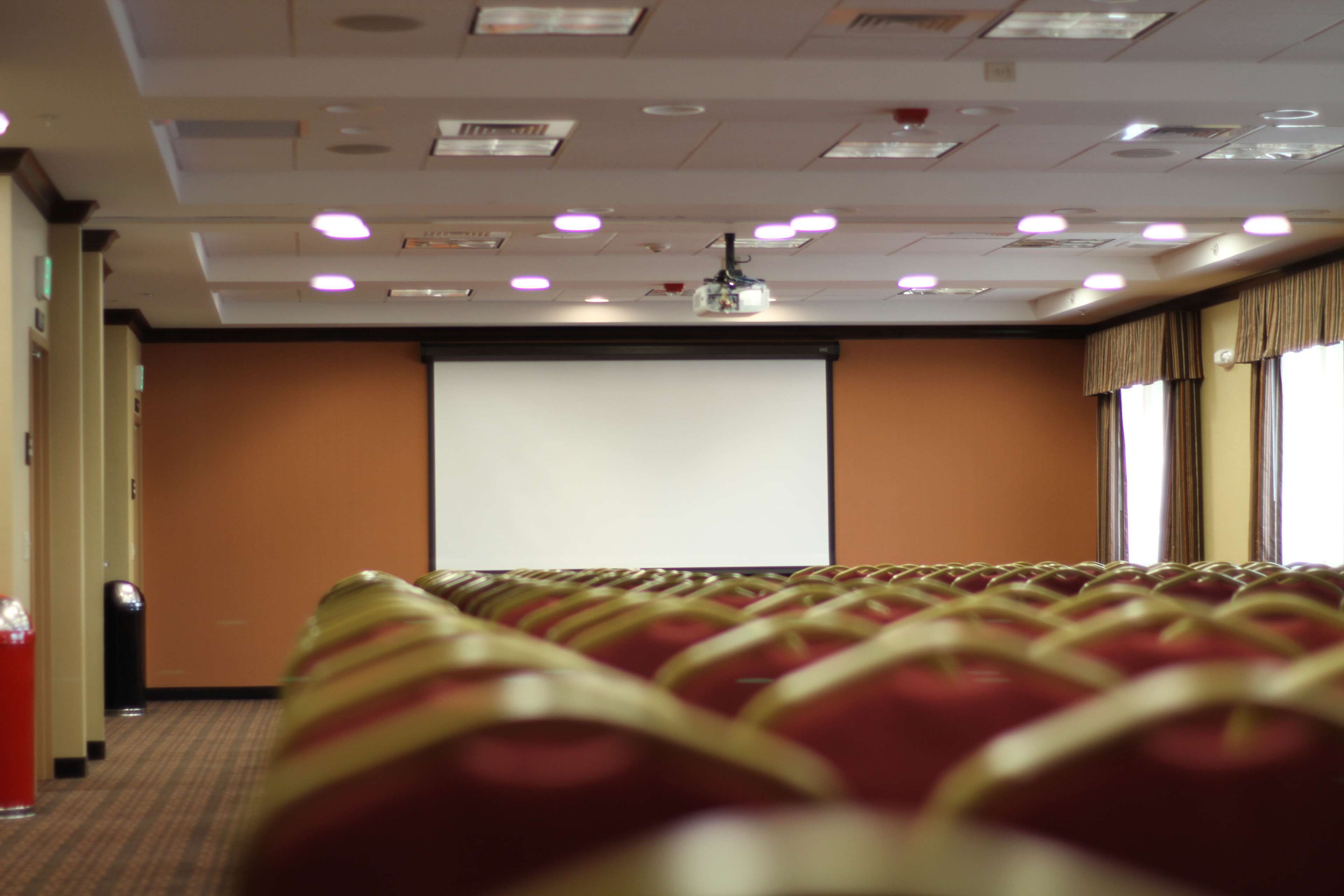 Un salón de conferencias con filas de sillas, una pantalla de proyector e iluminación empotrada en el techo.