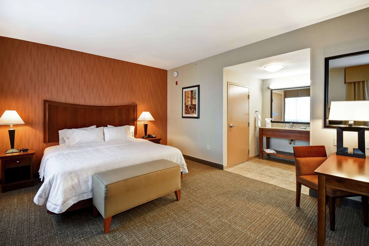 A hotel room with a bed, bedside tables, a desk, a chair, a mirror, and a bathroom door.