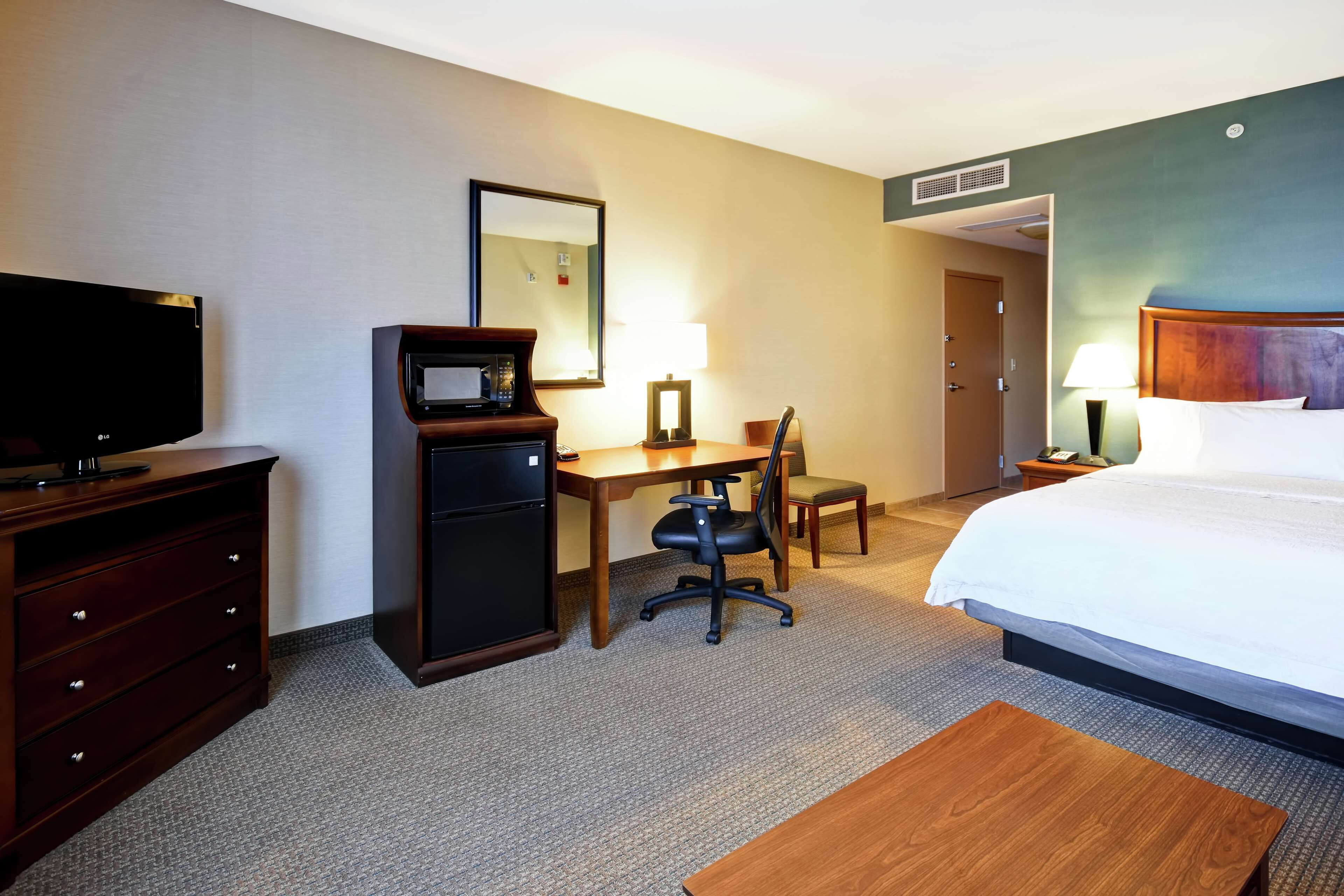 Quarto de hotel com cama, escrivaninha, cadeira, televisão e frigobar.