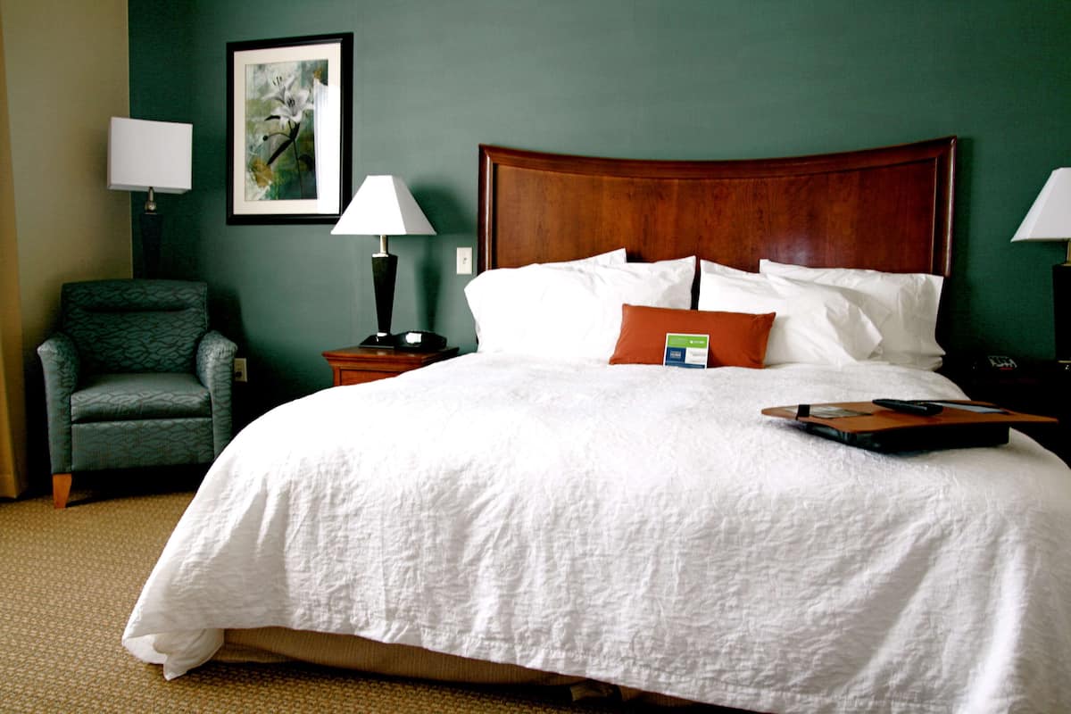 A hotel room with a large bed, a green armchair, two bedside tables with lamps, and a framed picture on the wall.