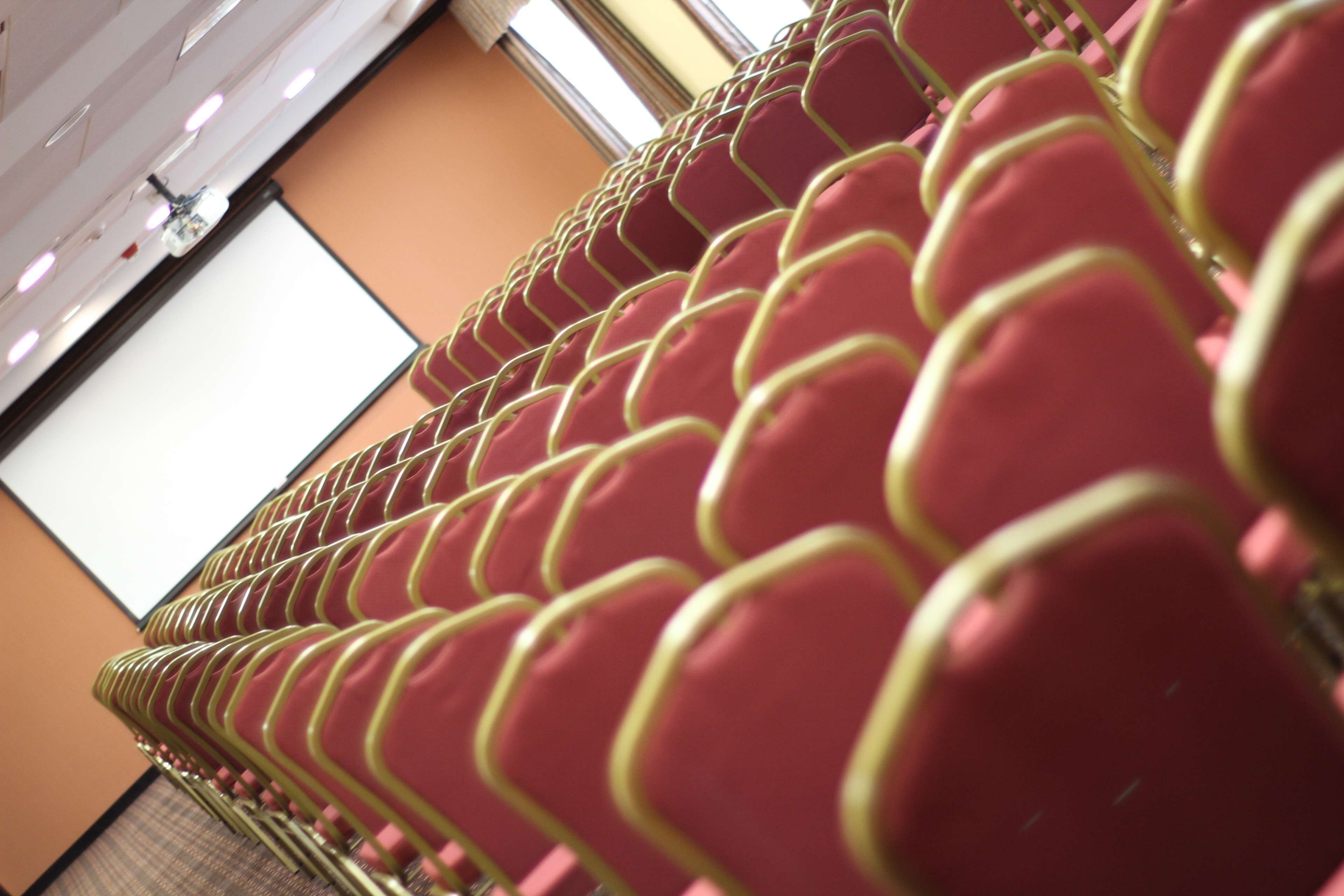 Rows of red chairs with yellow trim, arranged in a venue with a projection screen in the background.