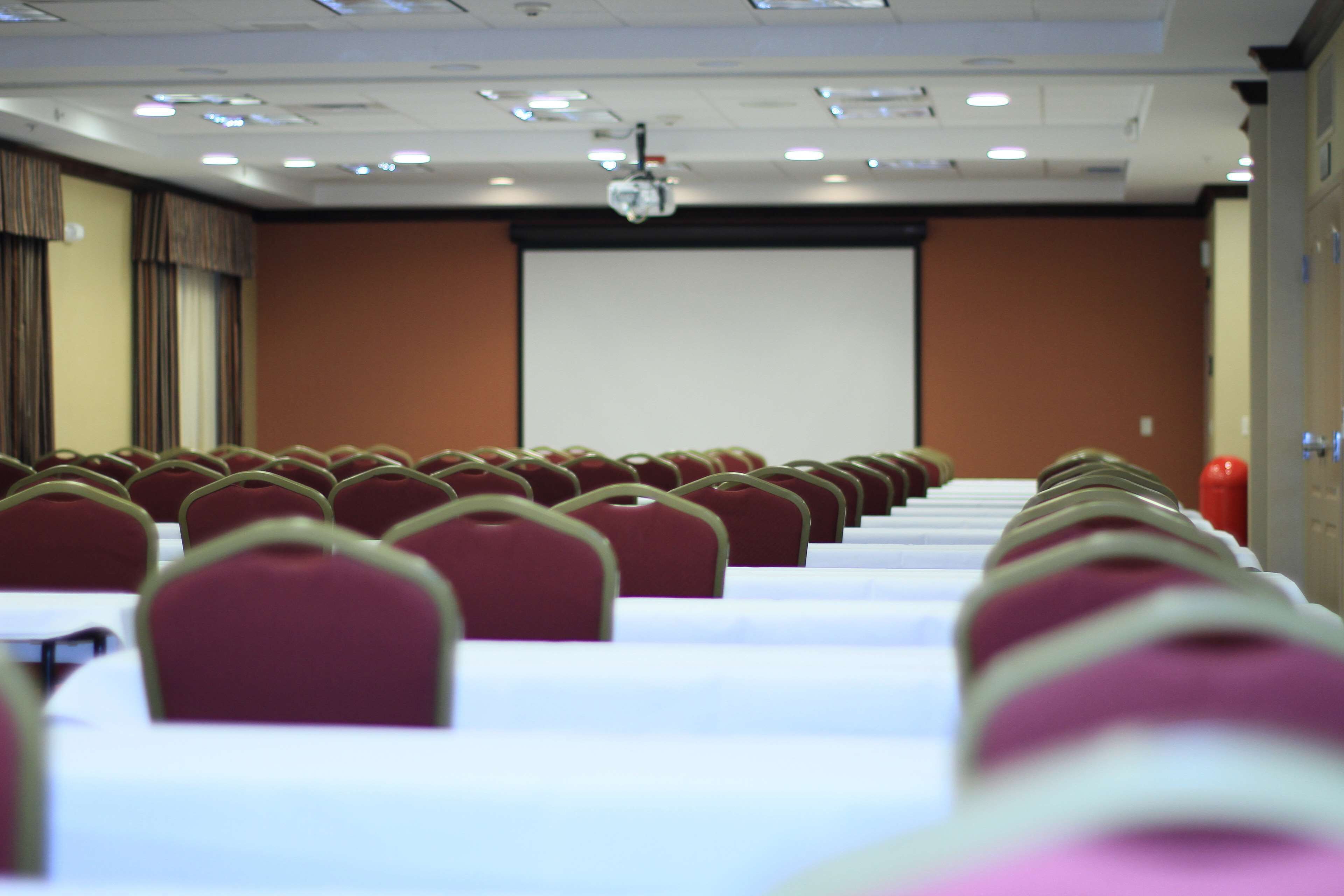 A conference room with rows of red chairs and white tables, a projector screen, and a fire extinguisher.