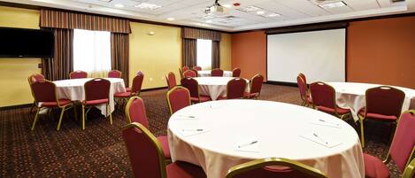 A conference room with multiple tables and chairs, a projector screen, and a large window with curtains.