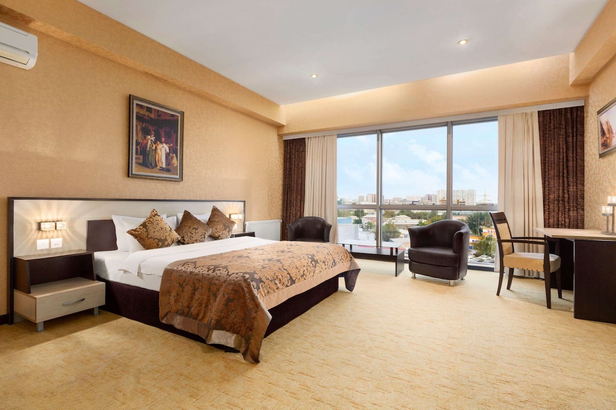Ein Hotelzimmer mit einem großen Bett, einem Schreibtisch, einem Stuhl und Blick auf die Stadt durch große Fenster.
