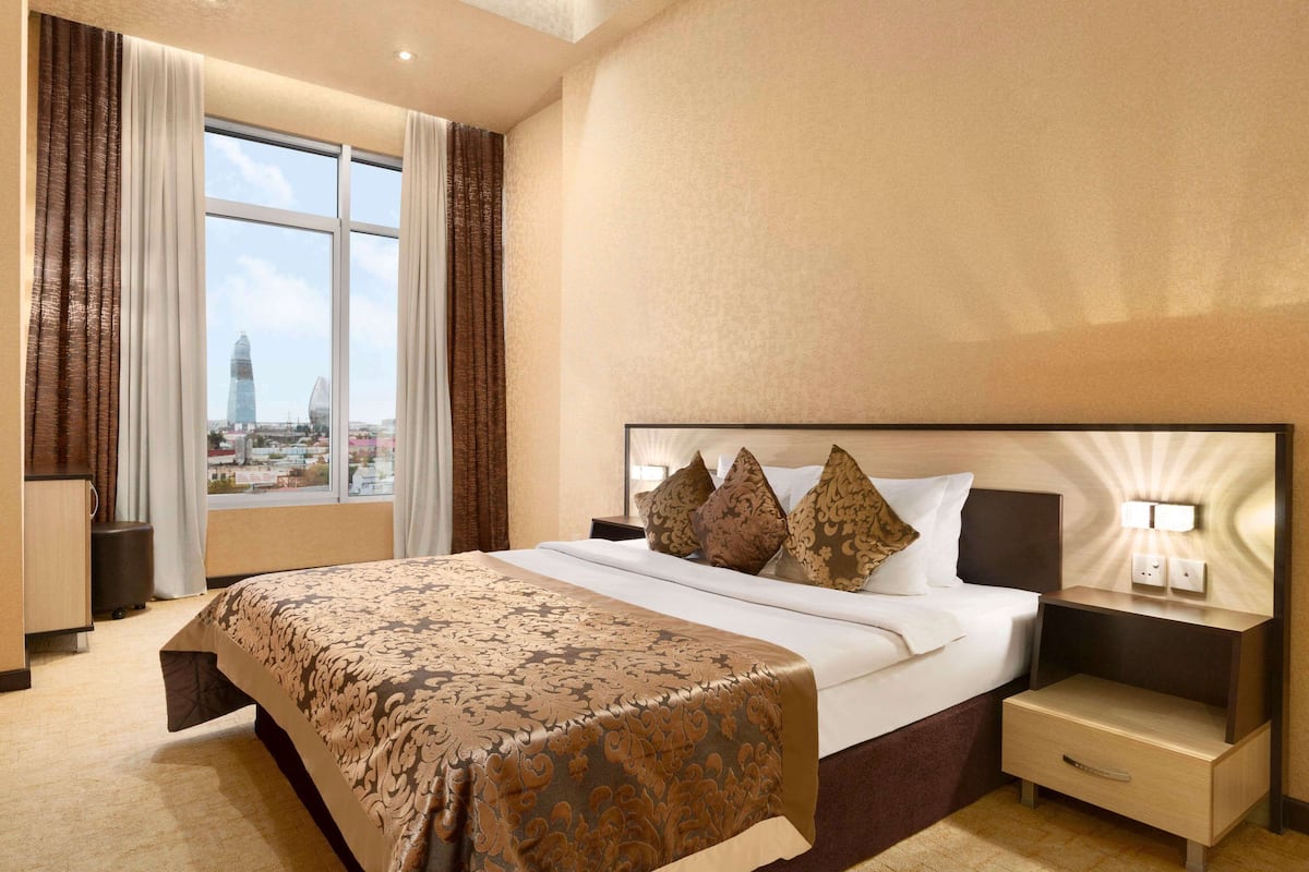 Ein Hotelzimmer mit einem großen Bett, einem Nachttisch, einer Schublade, einem Fenster mit Vorhängen und Blick auf die Stadt.