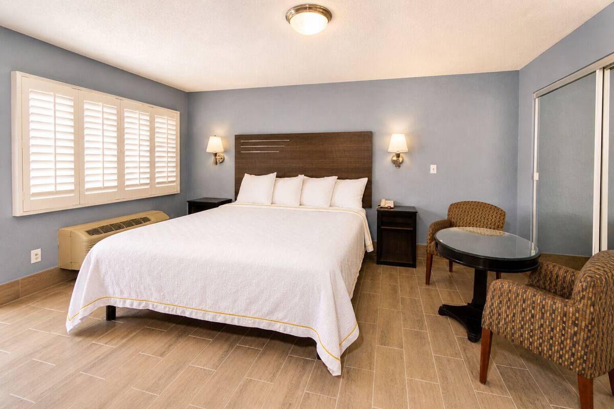 Una habitación de hotel con una cama grande, mesitas de noche, una silla, una pequeña mesa y una ventana con persianas.