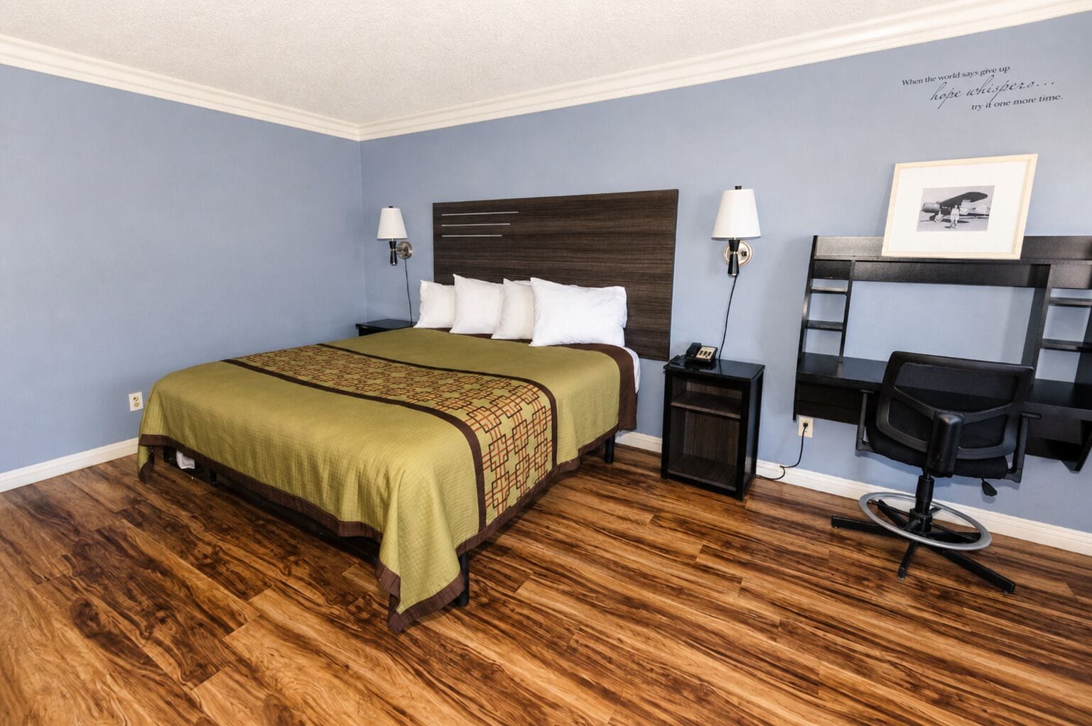 A hotel room with a bed, bedside lamps, a desk with a chair, and a motivational quote on the wall.