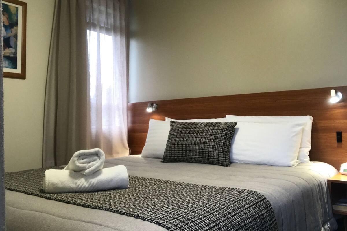 A neatly made bed with a grey and black patterned bedspread, white pillows, and a folded white towel on the side.