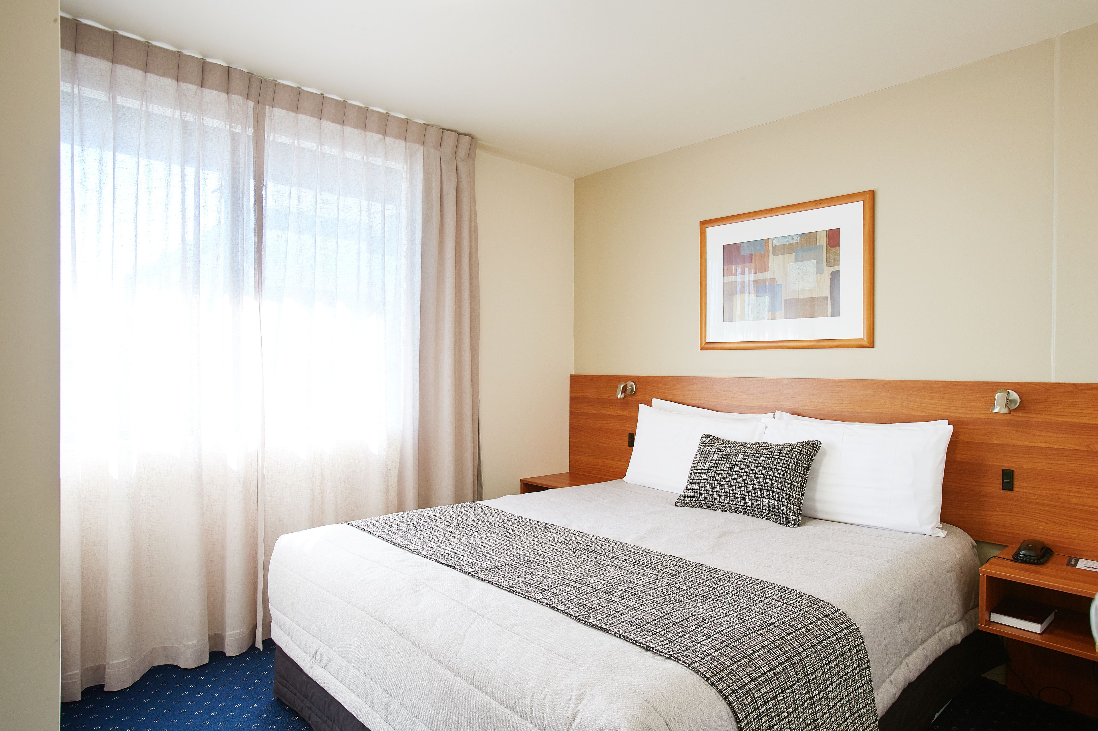 A hotel room with a large bed, a wooden headboard, a bedside table with a lamp, a framed picture on the wall, and sheer curtains on the window.
