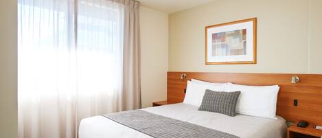 Una habitación de hotel con una cama grande, cabecera de madera, mesita de noche con lámpara, un cuadro enmarcado en la pared y cortinas translúcidas en la ventana.