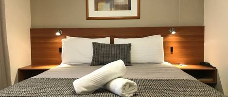 A neatly made bed with white and patterned pillows, a folded towel, and a framed abstract artwork above it.