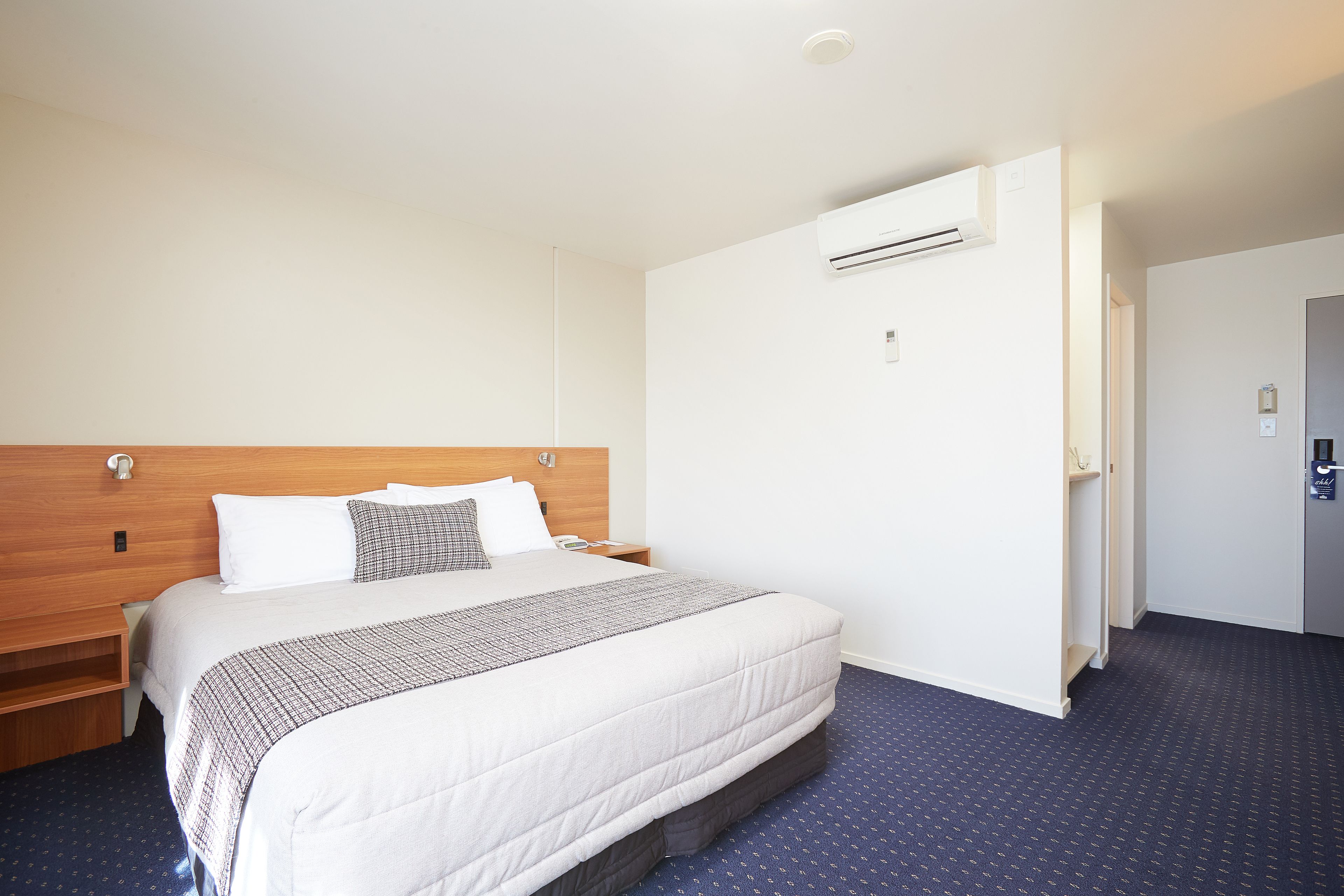 A hotel room with a large bed, a wooden headboard, a white wall, an air conditioning unit, and a blue carpet.