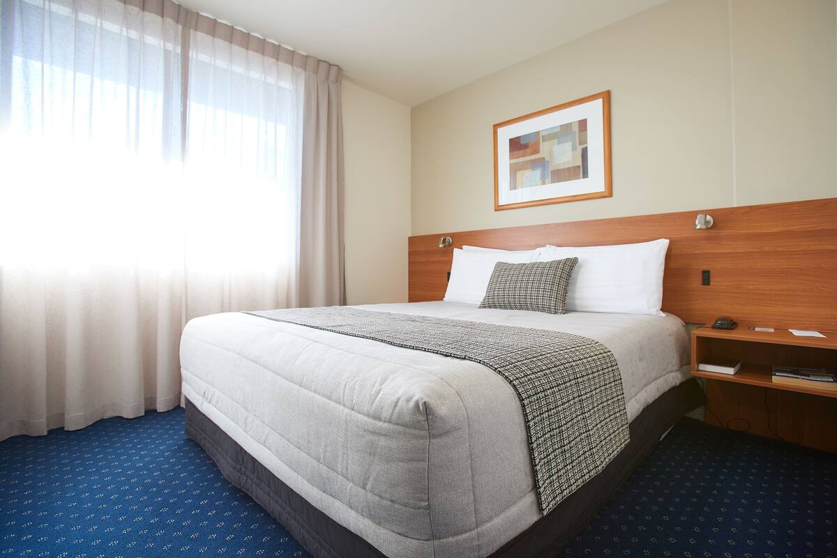 A hotel room with a large bed, a wooden headboard, a bedside table, a framed picture on the wall, and a window with curtains.