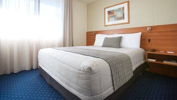 A hotel room with a large bed, a wooden headboard, a bedside table, a framed picture on the wall, and a window with curtains.