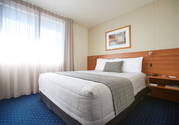 A hotel room with a large bed, a wooden headboard, a bedside table, a framed picture on the wall, and a window with curtains.