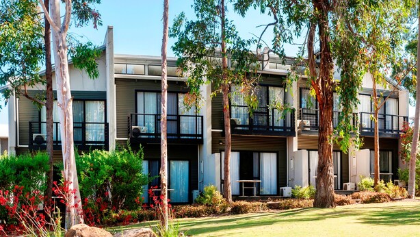 A modern multi-story building with balconies, surrounded by trees and landscaped gardens.