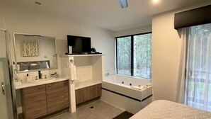 A modern hotel room with a large bed, a walk-in shower, a bathtub, a ceiling fan, a TV, and a window with a view of trees.