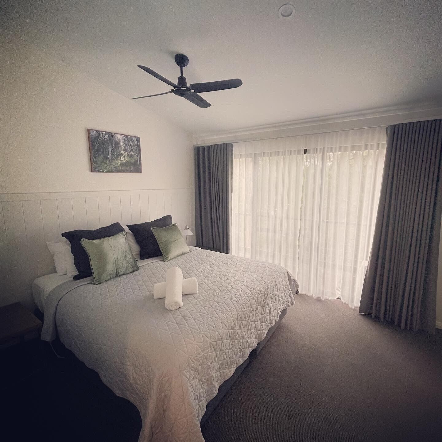 Una cama bien tendida con colcha, almohadas y una toalla enrollada. Se ve un ventilador de techo arriba.