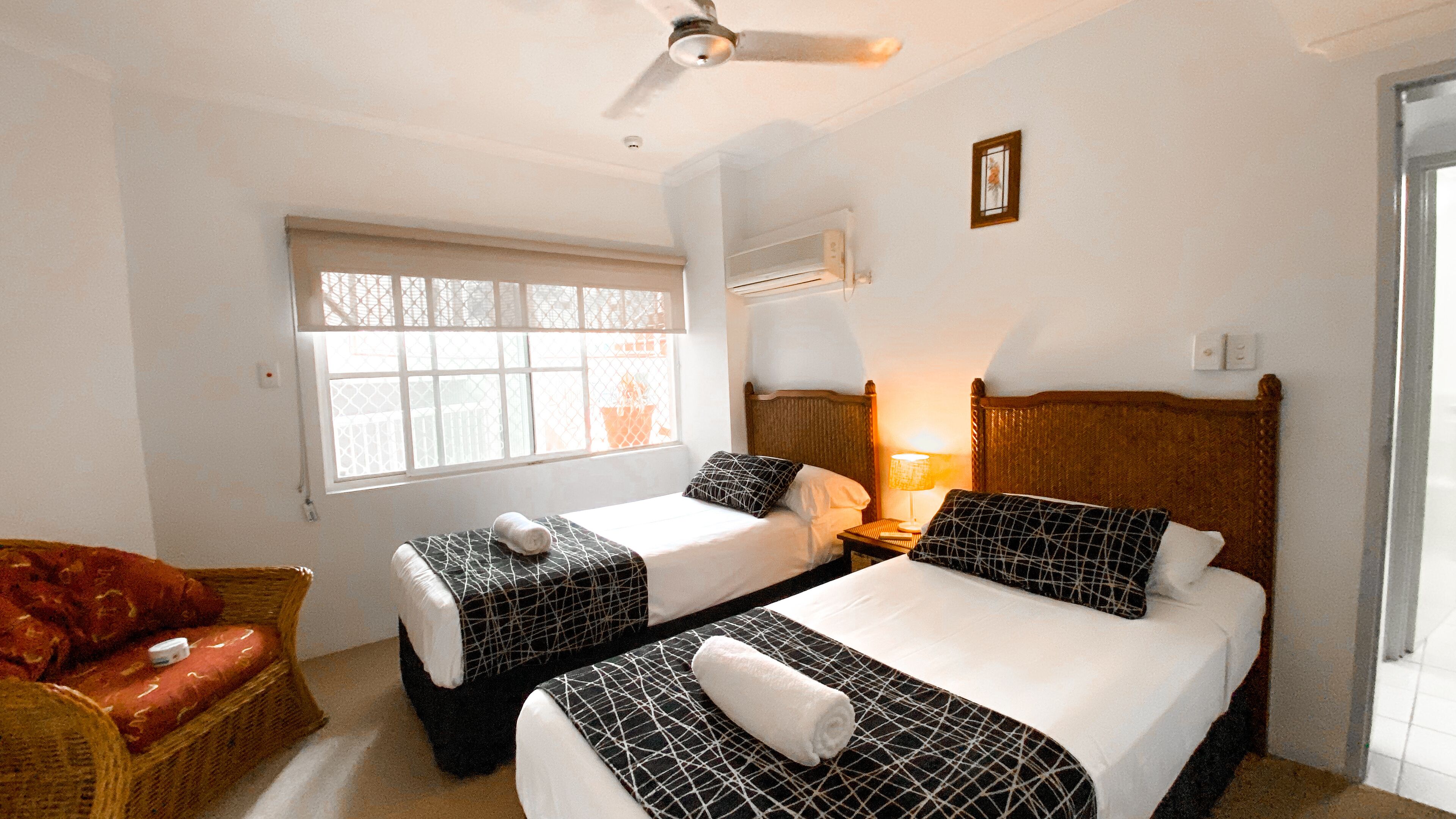 Habitación de hotel con dos camas, ventana con vistas, ventilador de techo y una silla de mimbre.