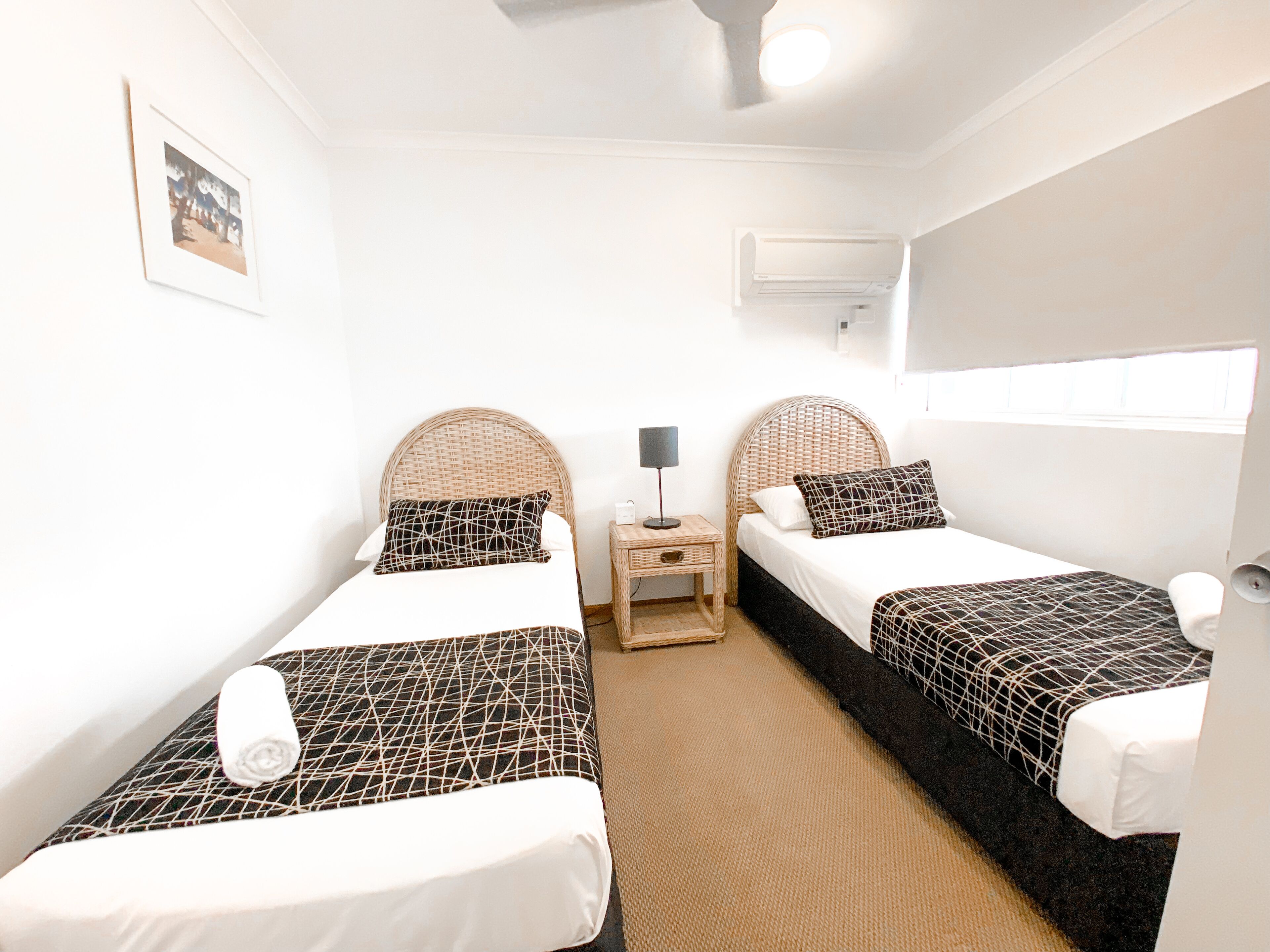 A hotel room with two single beds, a small bedside table with a lamp, a wall-mounted air conditioner, and a framed picture on the wall.