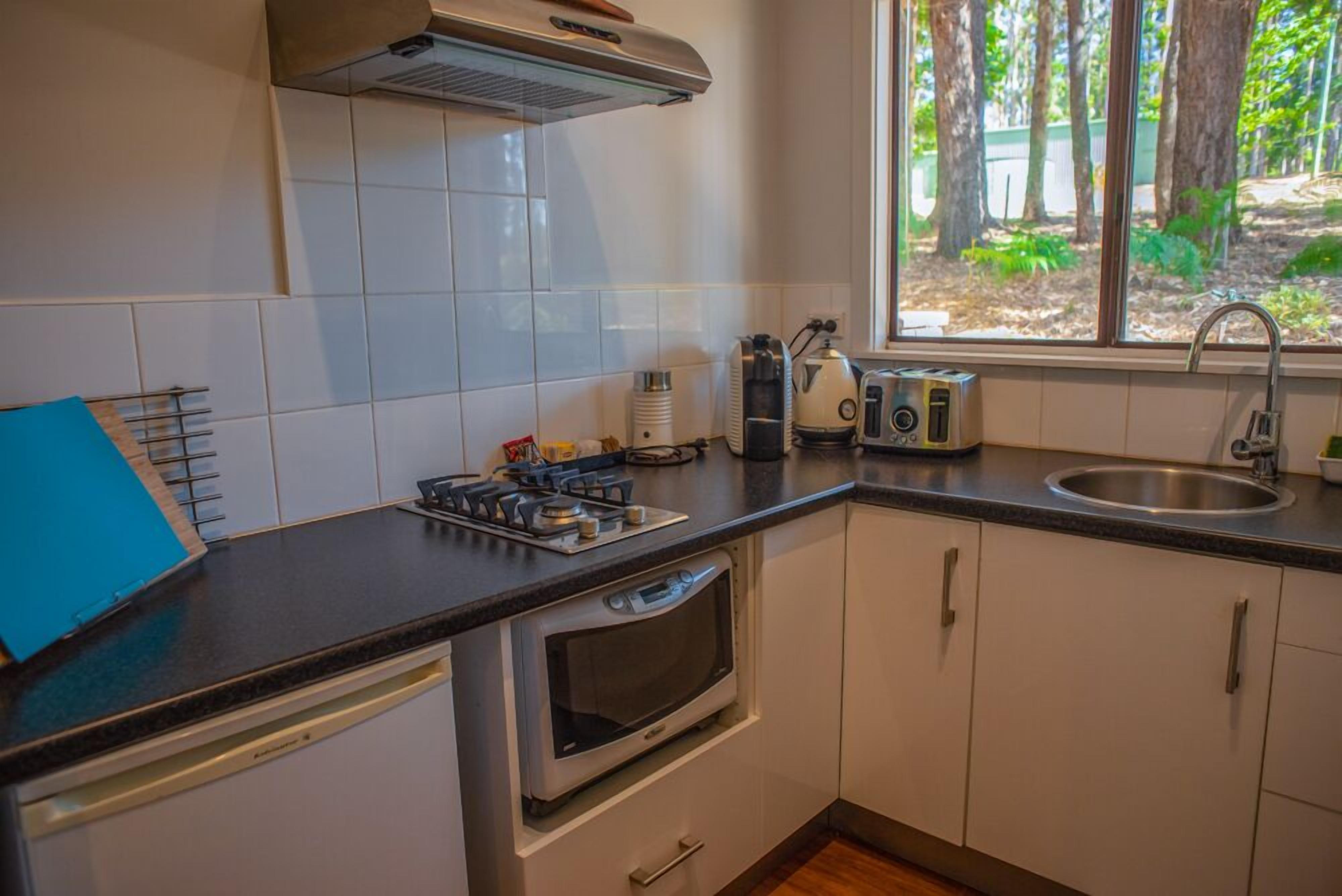 Une cuisine compacte avec des meubles blancs, un four encastré, une cuisinière à gaz et une fenêtre donnant sur les arbres.