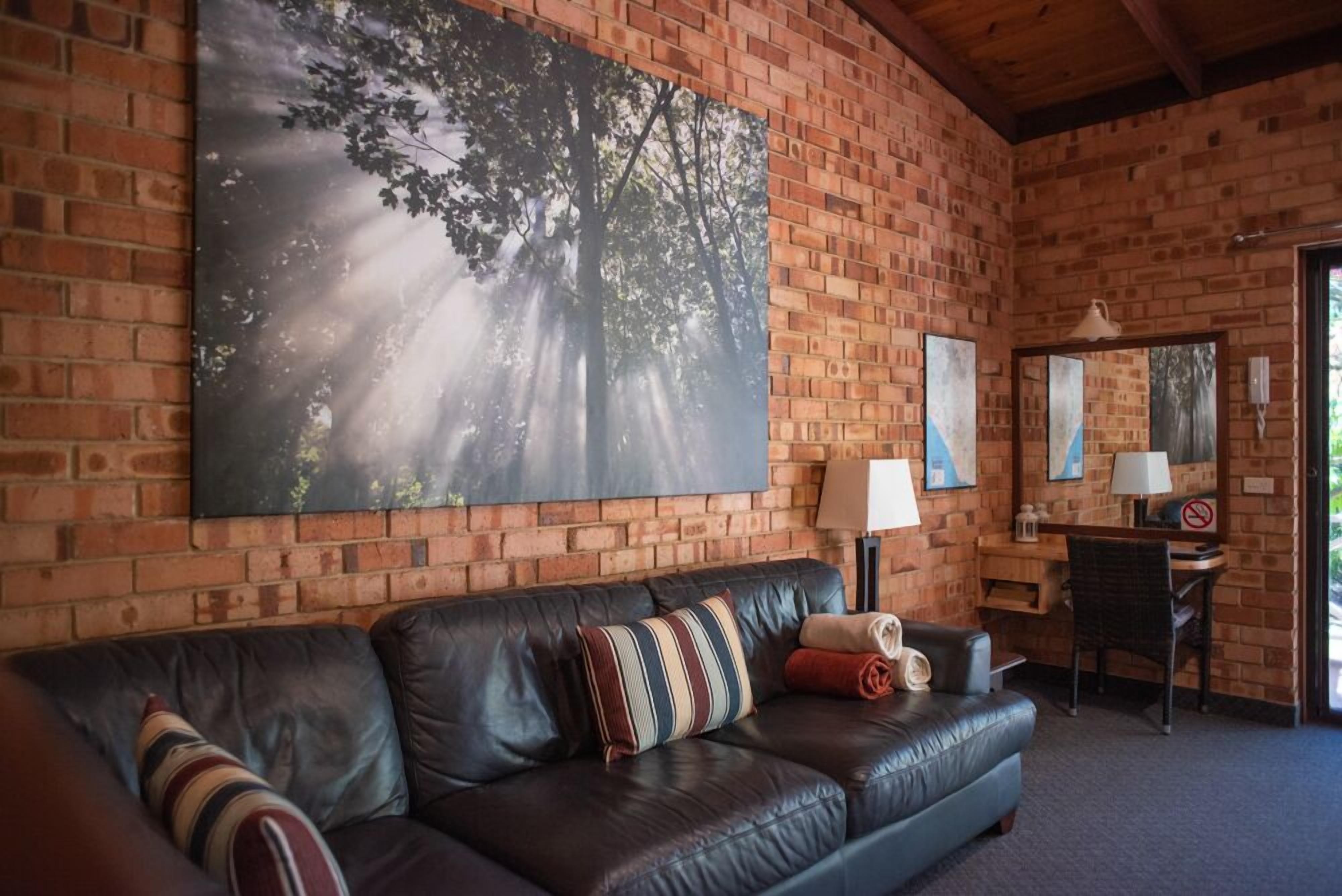 Et værelse med en murstensvæg, et stort maleri af træer, en lædersofa, et skrivebord med stol og en lampe.