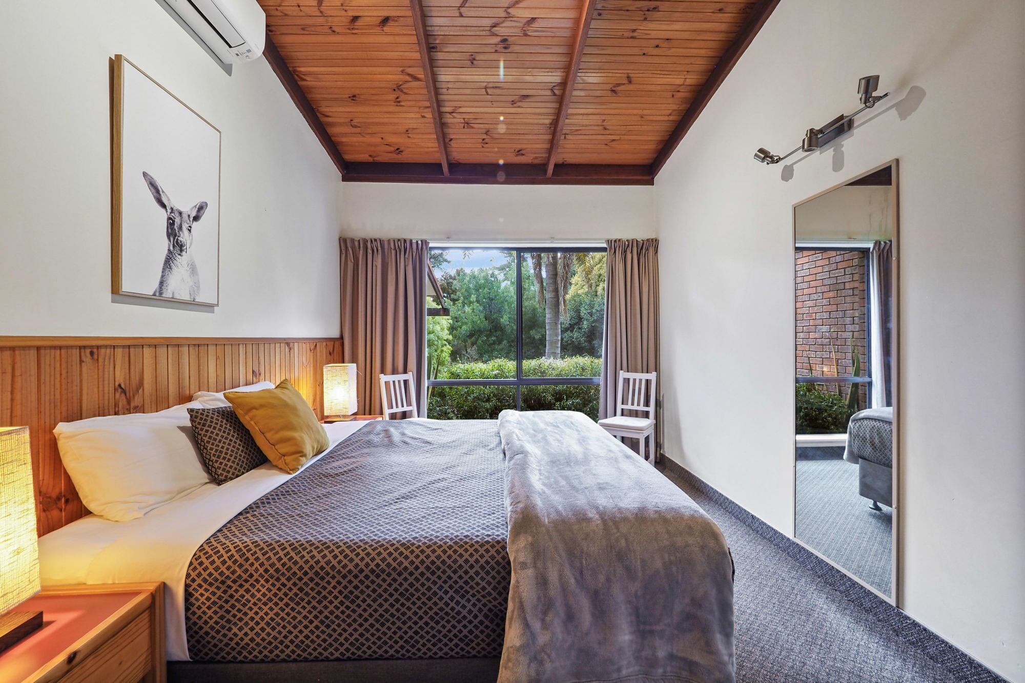 A bedroom with a large bed, two bedside tables with lamps, a headboard, a window with curtains, and a view of greenery outside.