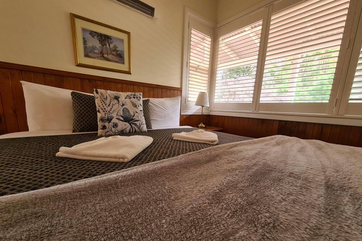 A neatly made bed with two pillows and a blanket, a bedside table with a lamp, and a window with blinds.
