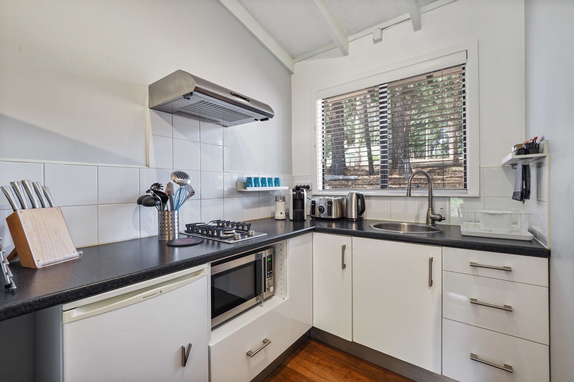 A modern kitchen with white cabinets, a black countertop, stainless steel appliances, and a window with blinds.