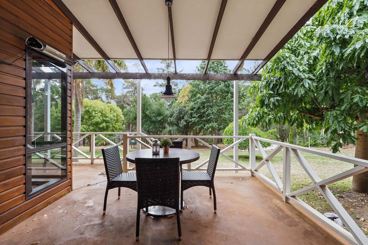 A covered patio with a round table and two chairs, a hanging light fixture, and a view of greenery.