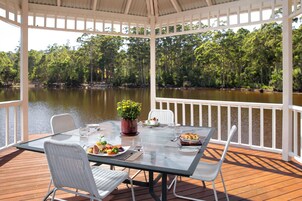 Un área de comedor al aire libre techada con una mesa lista para dos personas, con vistas a un lago rodeado de árboles.