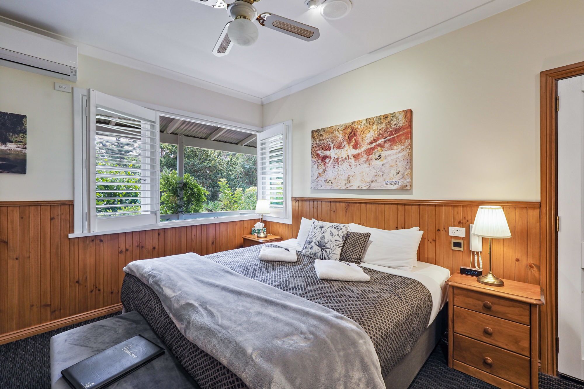 A hotel room with a bed, bedside table, nightstand, and a window with blinds.