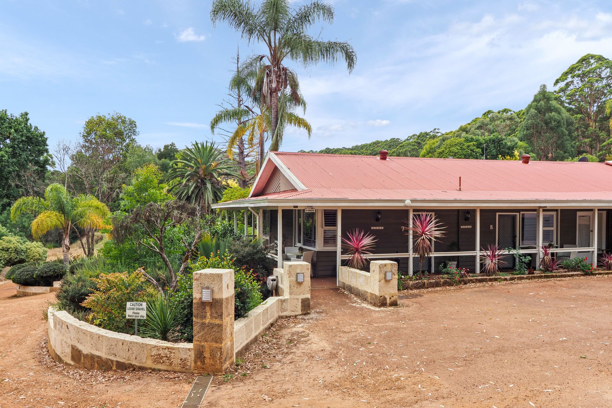 A two-story building with a red roof, surrounded by lush greenery and a dirt driveway.
