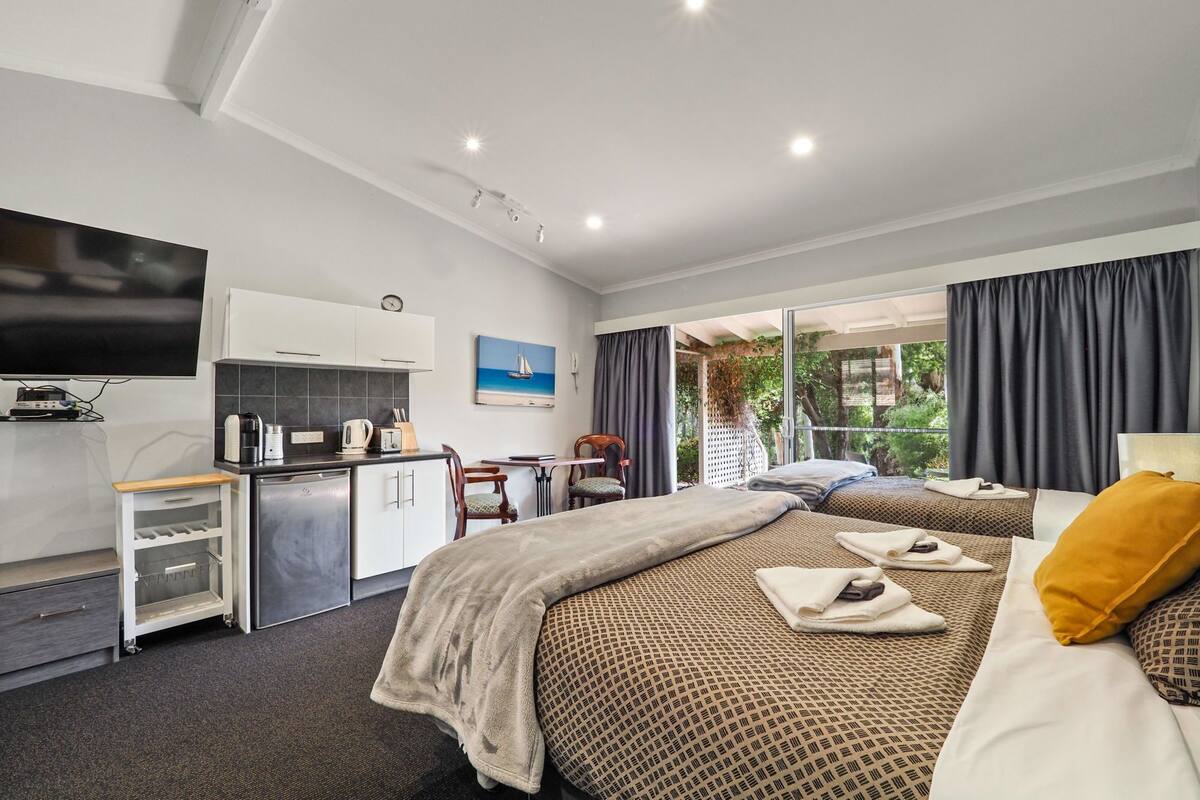 A modern hotel room with a kitchenette, a bed with a patterned duvet, a TV, and a view of greenery through the window.