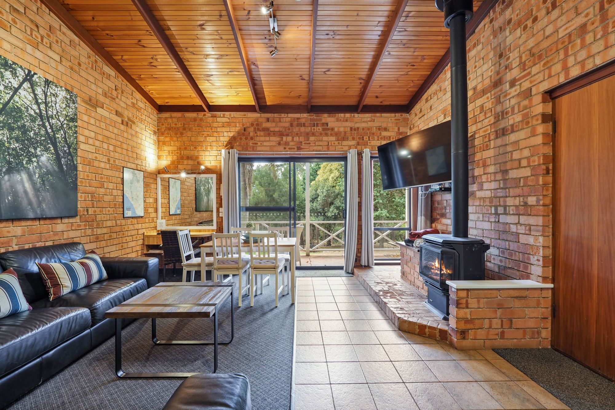 A cozy cabin interior with a fireplace, wooden ceiling, brick walls, a sofa, a dining table, and a TV.