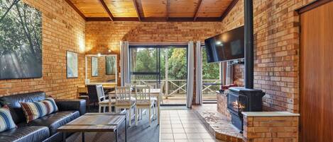 A cozy cabin interior with a fireplace, wooden ceiling, brick walls, a sofa, a dining table, and a TV.
