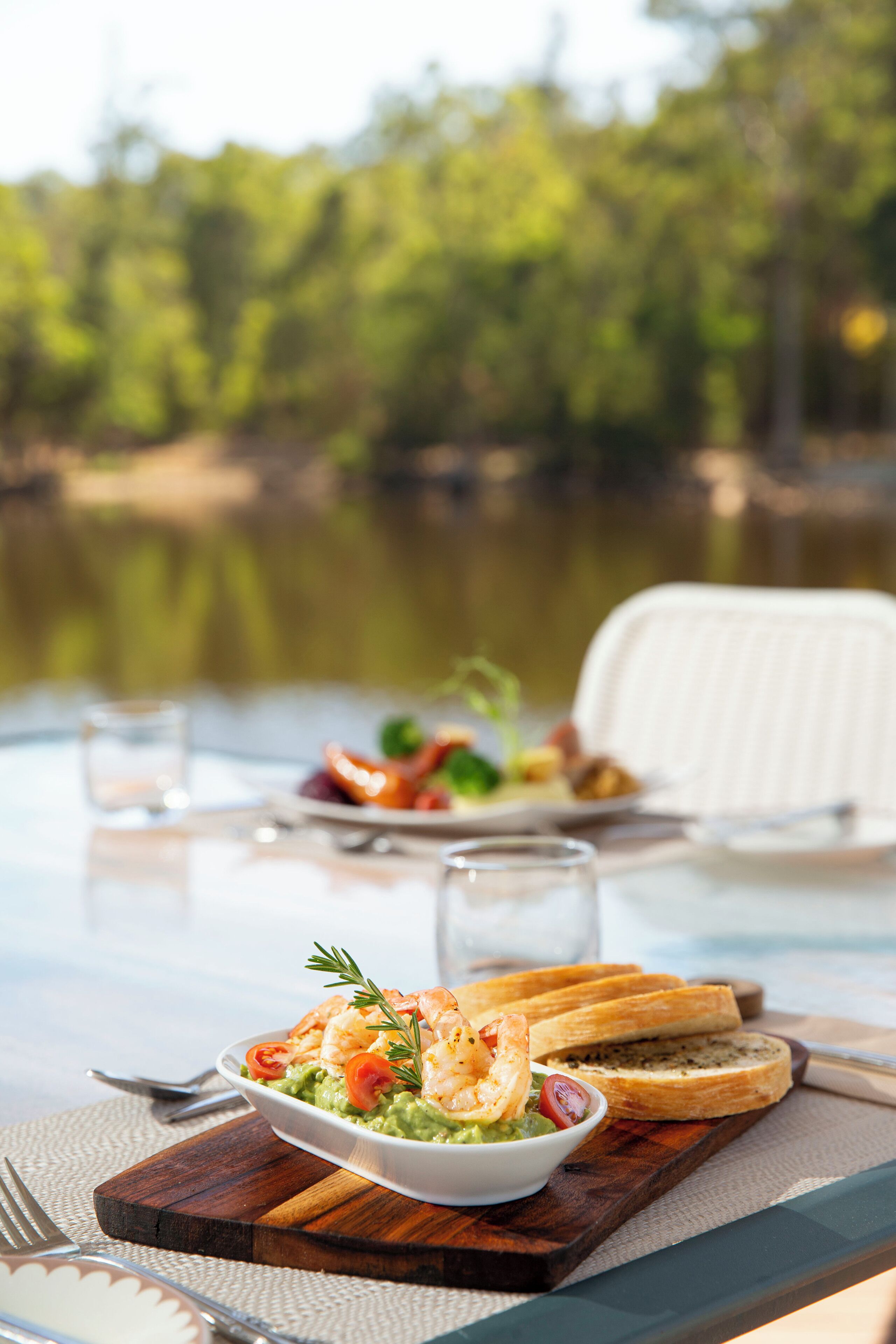A dining table with a seafood dish, bread, and a glass of water, set against a lakeside backdrop.