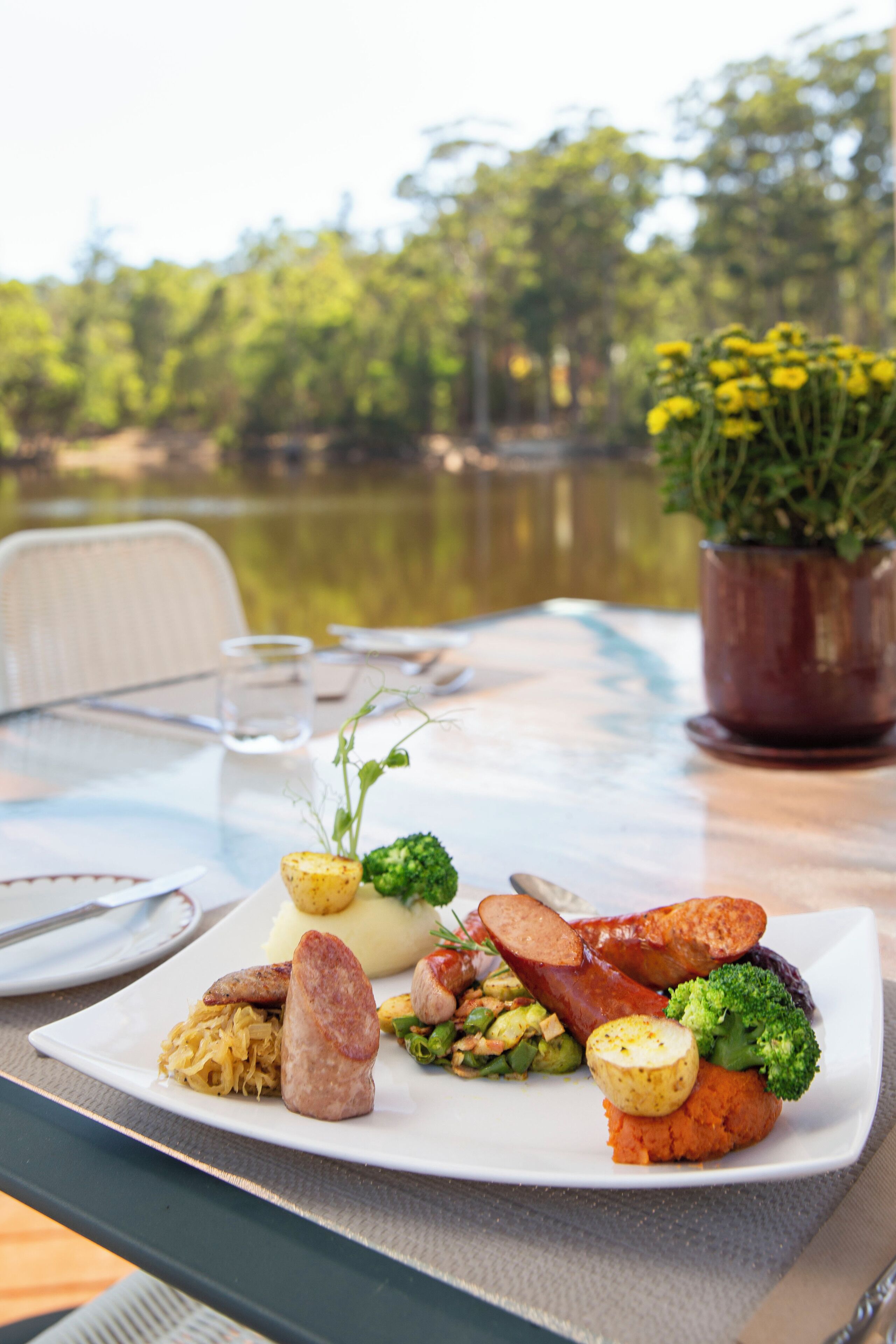 A dining table with a variety of dishes, including sausages, potatoes, and vegetables, set against a backdrop of a lake and greenery.