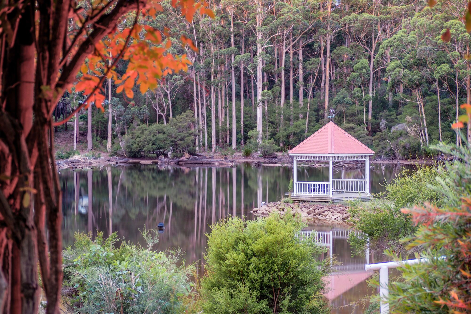 A gazebo with a red roof and white railings, surrounded by a calm lake and dense forest.