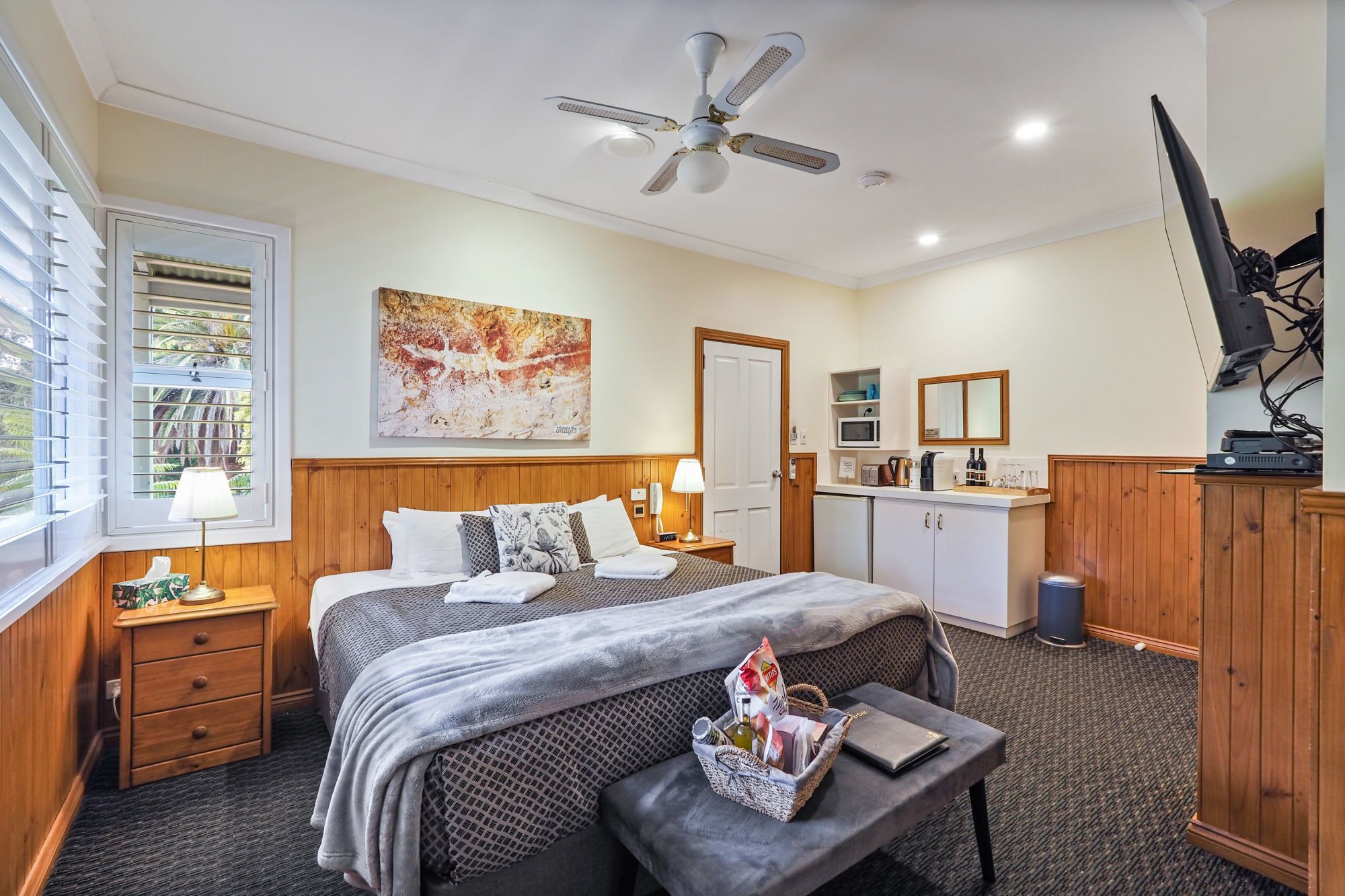 A hotel room with a bed, bedside table, lamp, artwork, kitchenette, and a television.