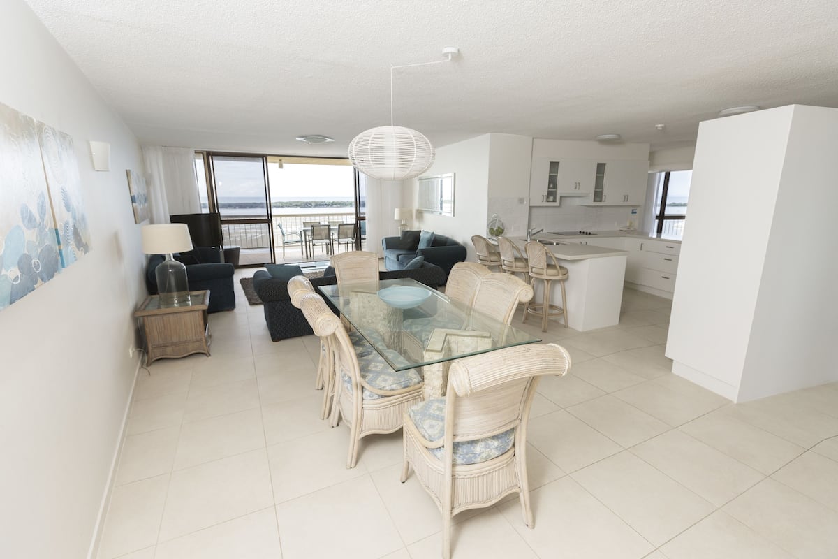 A modern living room with a glass dining table, wicker chairs, a sofa, and a view of the sea.