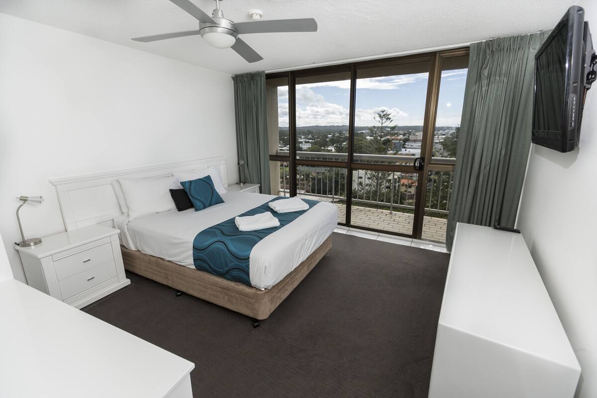 A modern hotel bedroom with a large bed, a bedside table, a nightstand, a ceiling fan, a TV mounted on the wall, and a balcony with a view of the city.
