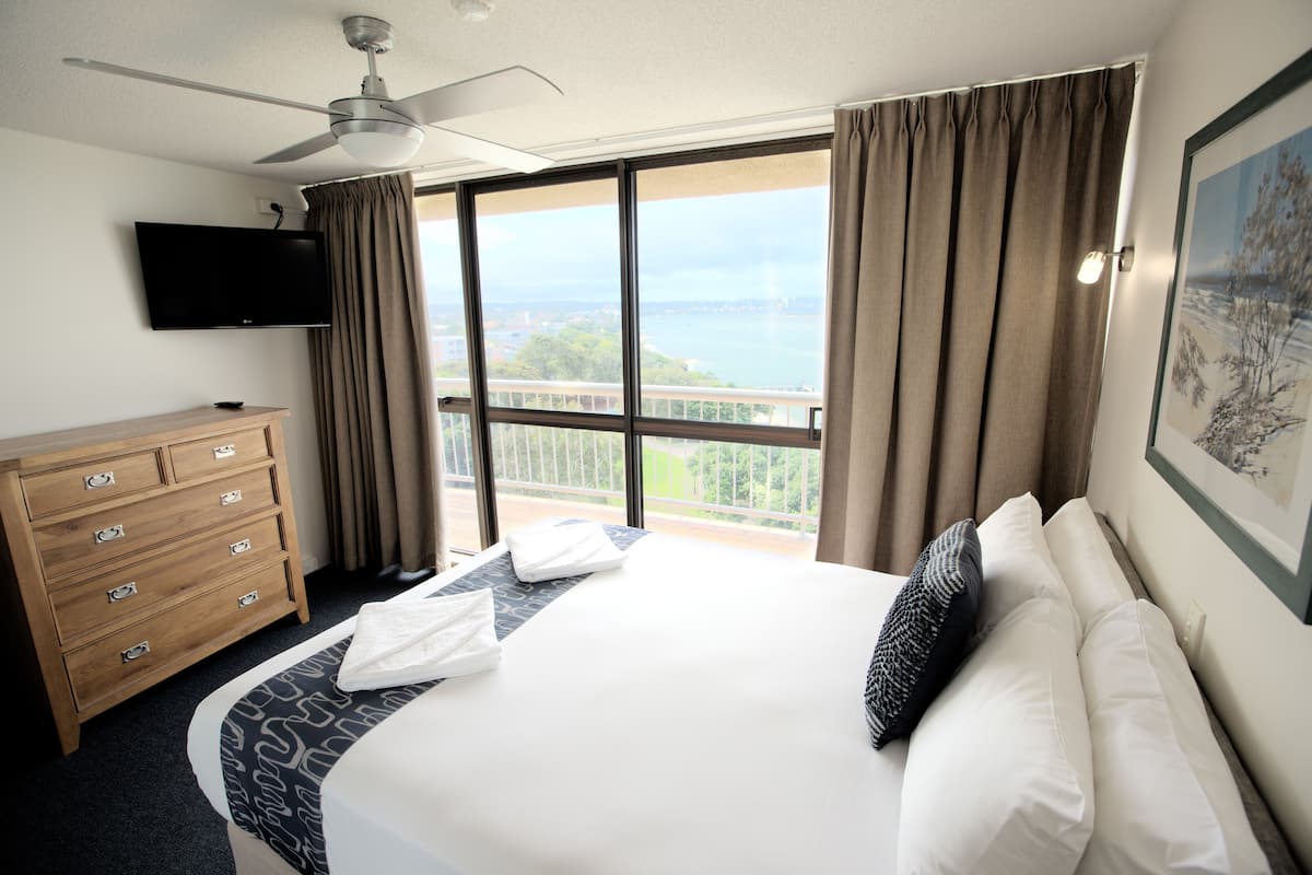 A hotel room with a bed, a wooden dresser, a flat-screen TV, and a balcony with a view of the city.