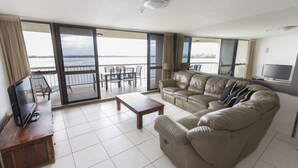 En rummelig opholdsstue med en lædersofa, et træbord og et fjernsyn på et træstel. Store skydedøre fører ud til en balkon med udsigt over vandet.