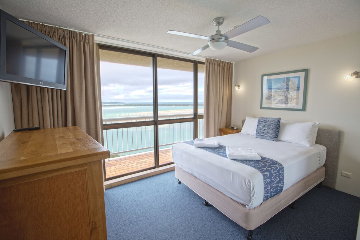 A hotel room with a bed, a TV, a ceiling fan, a large window with a view of the beach, and a wooden desk.