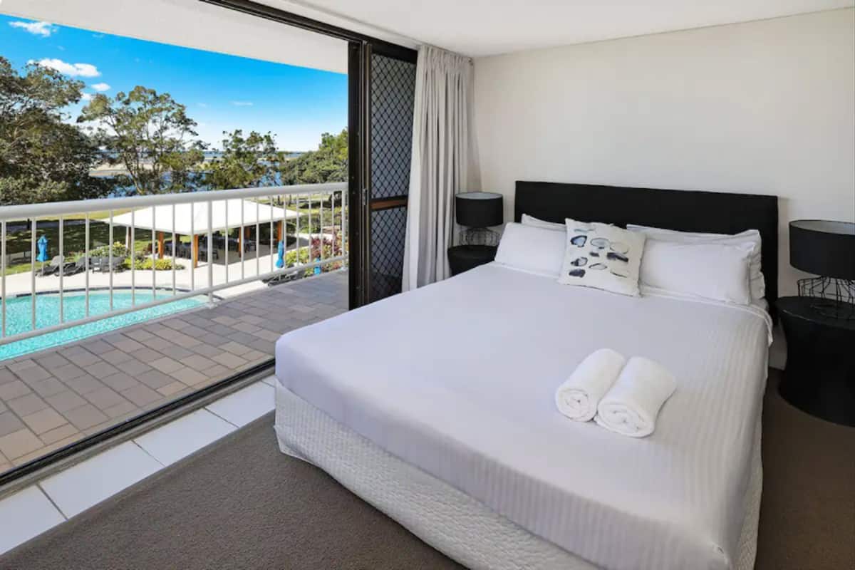 A hotel room with a large bed, two bedside tables, a view of a pool and outdoor area.